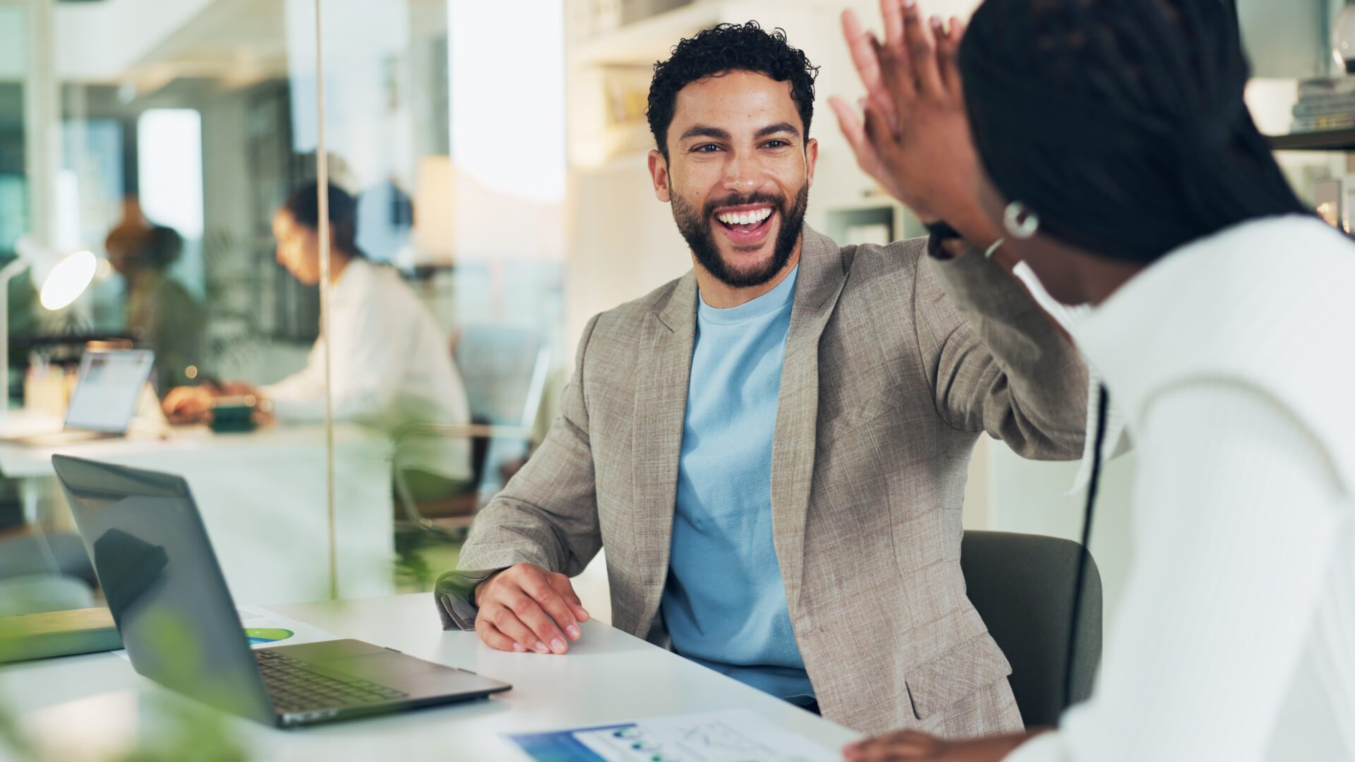 Two people seated at a desk in a modern office, raising their hands toward each other in a celebratory gesture. A laptop and printed documents are on the table, with other people working in the background.