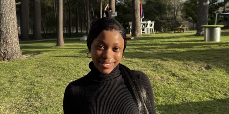 Person standing outdoors on grass with tall trees behind them, facing the camera in natural daylight.