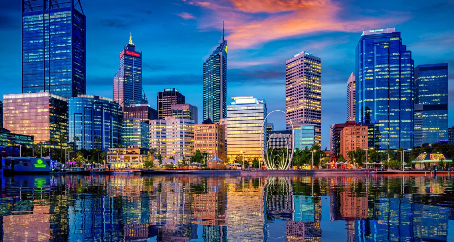 City skyline at dusk with illuminated high‑rise buildings reflected on calm water under a colourful evening sky.