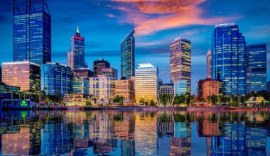 City skyline at dusk with illuminated high‑rise buildings reflected on calm water under a colourful evening sky.