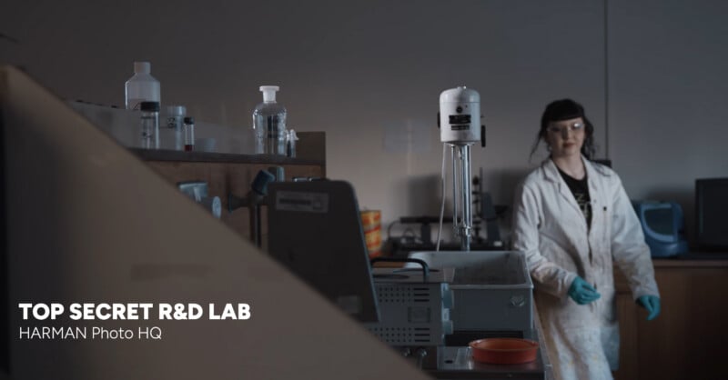 A scientist in a white lab coat and gloves stands in a dimly lit laboratory with various lab equipment and bottles on the counters. Text in the lower left corner reads, "TOP SECRET R&D LAB HARMAN Photo HQ.