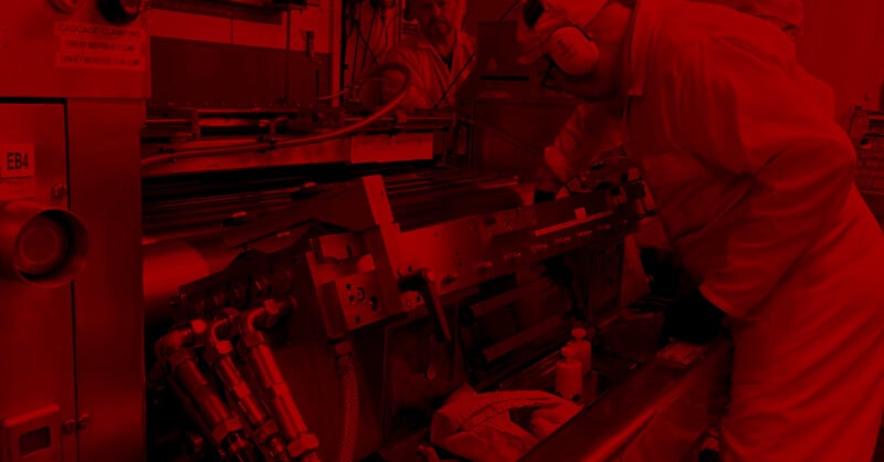 Two workers in protective clothing and ear protection operate and inspect a large industrial machine in a factory setting, with the entire image tinted red.