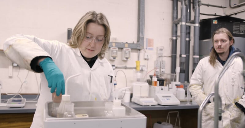 Two people in lab coats and safety glasses are in a laboratory. One person is using a piece of equipment with bottles, while the other stands nearby, observing. The lab has scientific instruments and cables in the background.