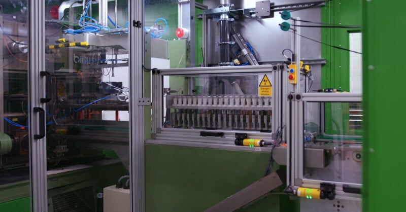 Automated industrial machine with metal framework and glass panels, filled with tubes and wires, assembling or inspecting small cylindrical components. Safety warnings and control buttons are visible on the equipment.