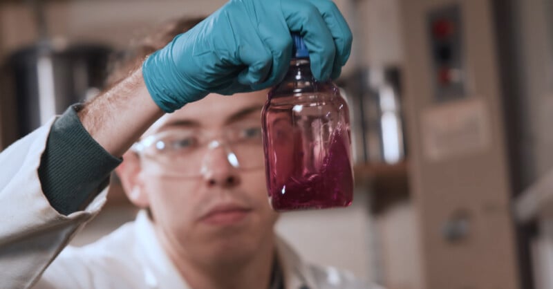 A scientist wearing safety glasses and teal gloves holds up a clear bottle containing a purple liquid, examining it closely in a laboratory setting.