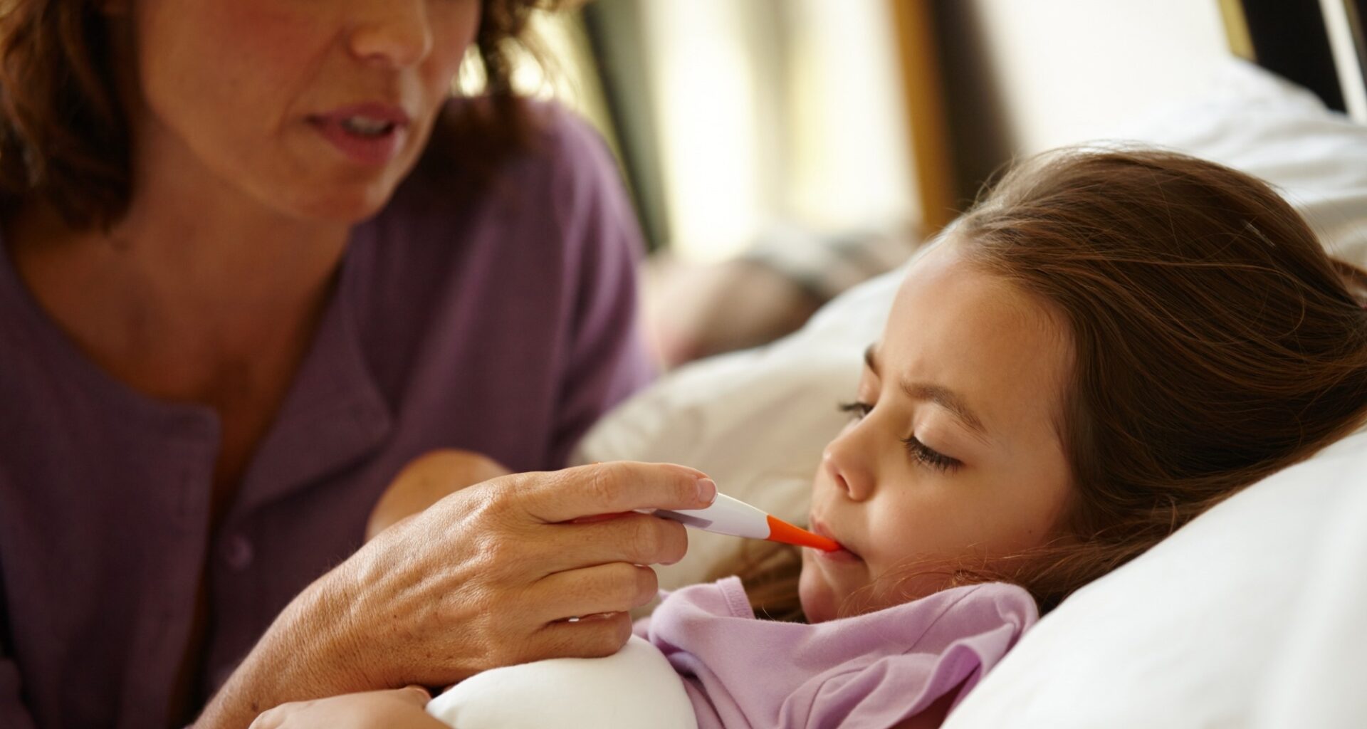 mother and daughter with thermometer by bed, monitor fever and symptoms concern for wellness check