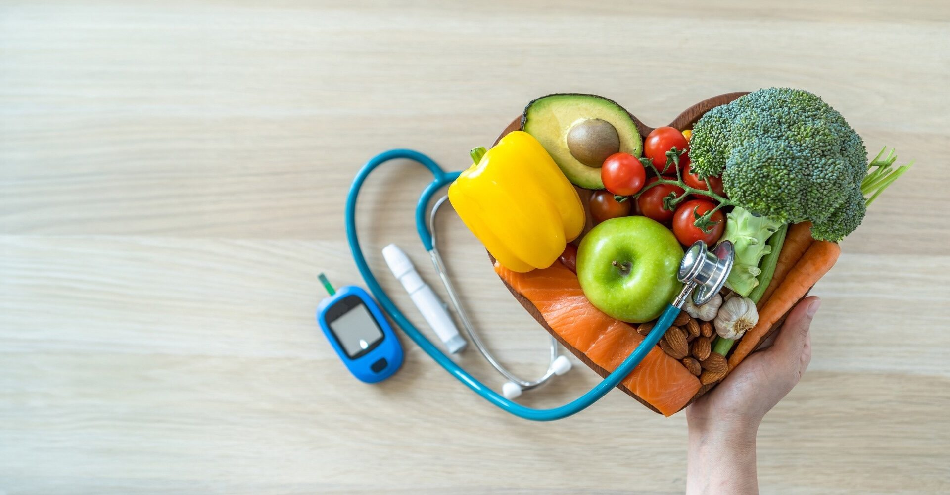 A hand holds a heart-shaped bowl filled with fresh healthy foods - including vegetables, fruit, fish, and nuts - next to a glucose meter and stethoscope, symbolizing diet and heart health management in diabetes.