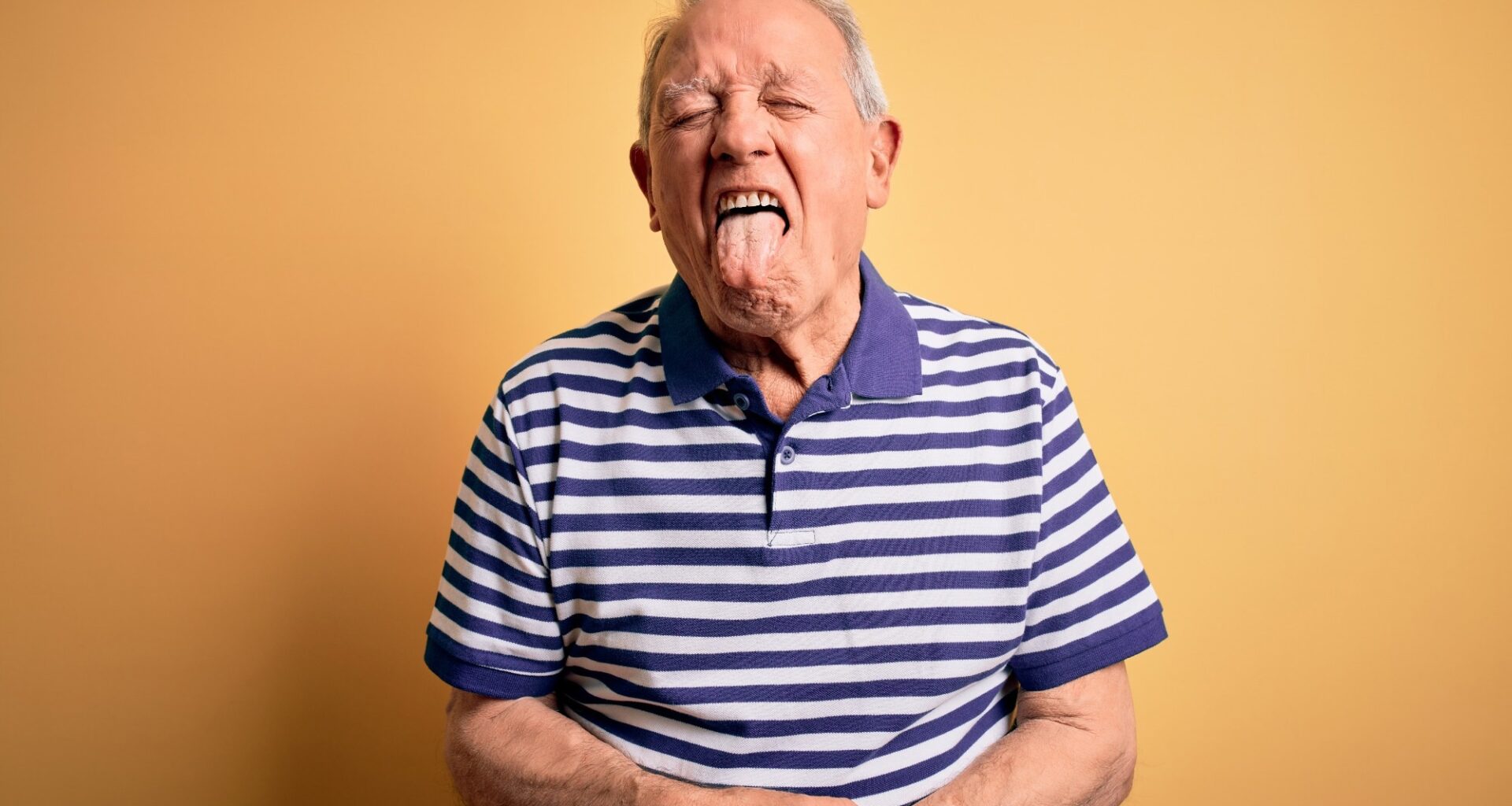 Grey haired senior man wearing casual navy striped t-shirt standing over yellow background sticking tongue out happy with funny expression.