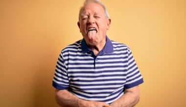 Grey haired senior man wearing casual navy striped t-shirt standing over yellow background sticking tongue out happy with funny expression.