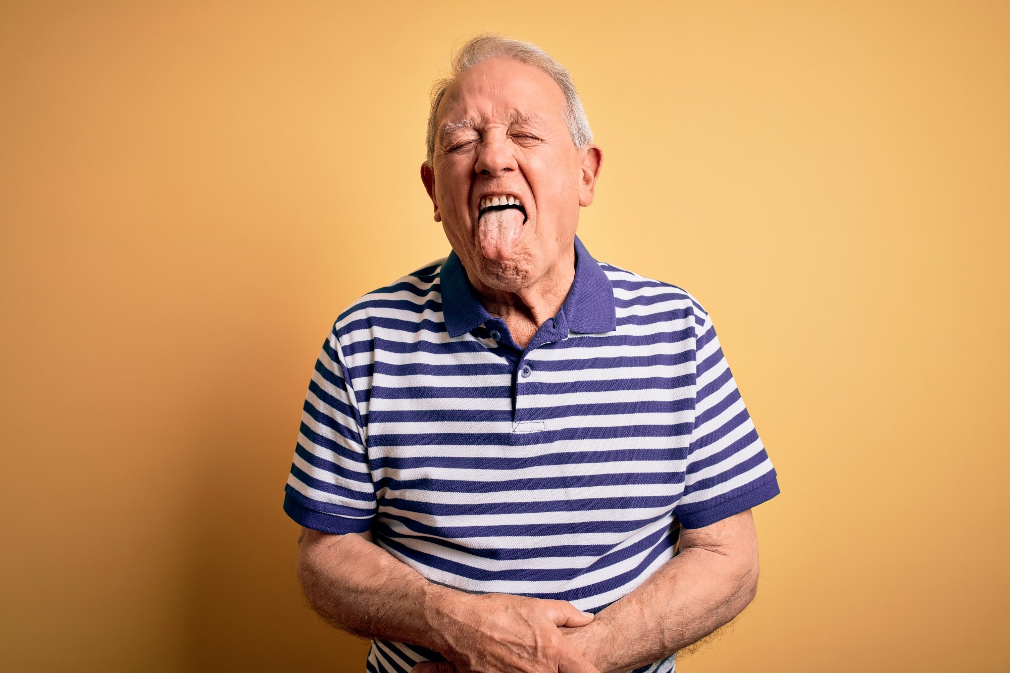 Grey haired senior man wearing casual navy striped t-shirt standing over yellow background sticking tongue out happy with funny expression.