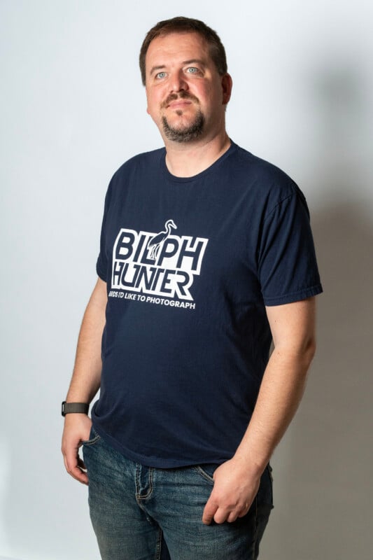 A man with short hair and a beard stands against a plain background, wearing a navy blue "Bird Hunter" T-shirt and jeans, with one hand in his pocket and a relaxed expression.