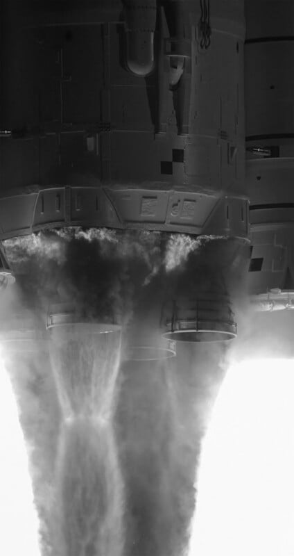 Close-up, black and white image of a rocket engine during liftoff, showing intense plumes of smoke and vapor emerging from the nozzles against a bright background.