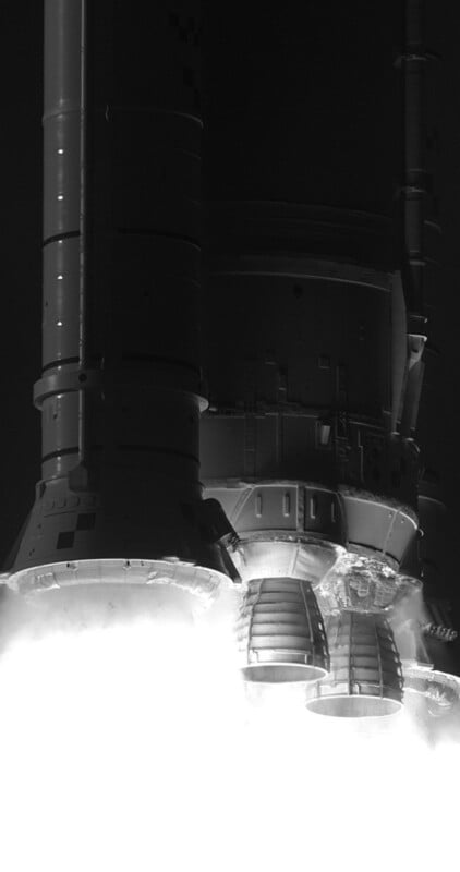 Close-up, black-and-white view of a rocket’s engines at liftoff, with bright flames and smoke pouring out beneath, highlighting the details of the engine nozzles and lower section of the spacecraft.