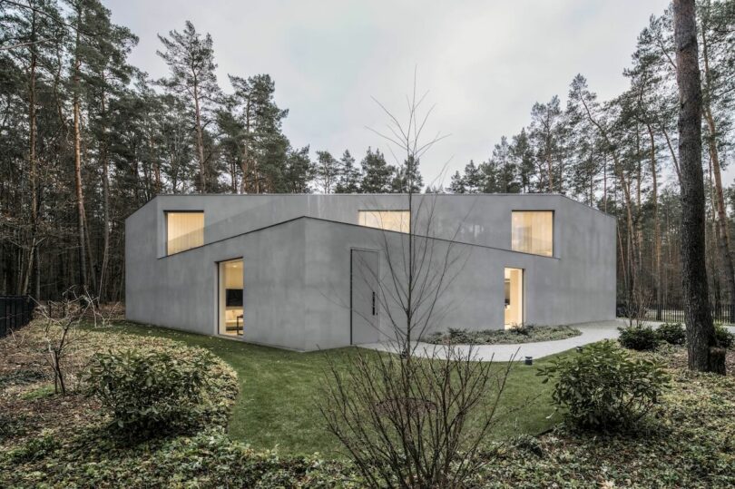 Modern angular grey concrete house by KWK Promes with large windows, surrounded by grass, shrubs, and tall pine trees in a wooded area.