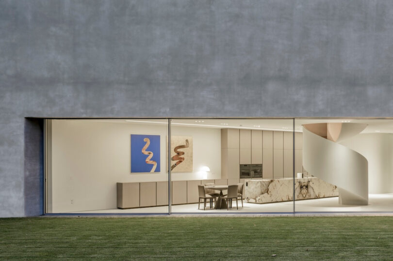 A modern kitchen and dining area designed by KWK Promes features minimalist decor, marble surfaces, a spiral staircase, and expansive floor-to-ceiling windows overlooking a lush lawn.