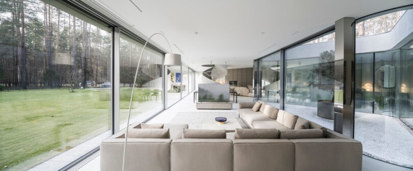 Modern living room designed by KWK Promes, featuring a large beige sectional sofa, glass walls, and views of a green lawn and forest. Minimalist decor and abundant natural light fill the space.