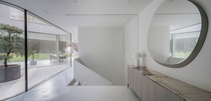 Minimalist modern interior by KWK Promes featuring large glass windows, a marble-topped console, round mirror, and white lamp, with an outdoor garden visible through the sleek windows.