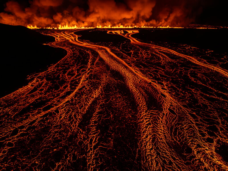 Molten lava flows down a dark landscape, glowing bright orange and red. In the background, a line of fire burns intensely, with thick smoke billowing upwards into the night sky.