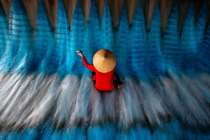 A person wearing a conical hat and red shirt sits surrounded by blue fishing nets, arranging or repairing them, viewed from above.