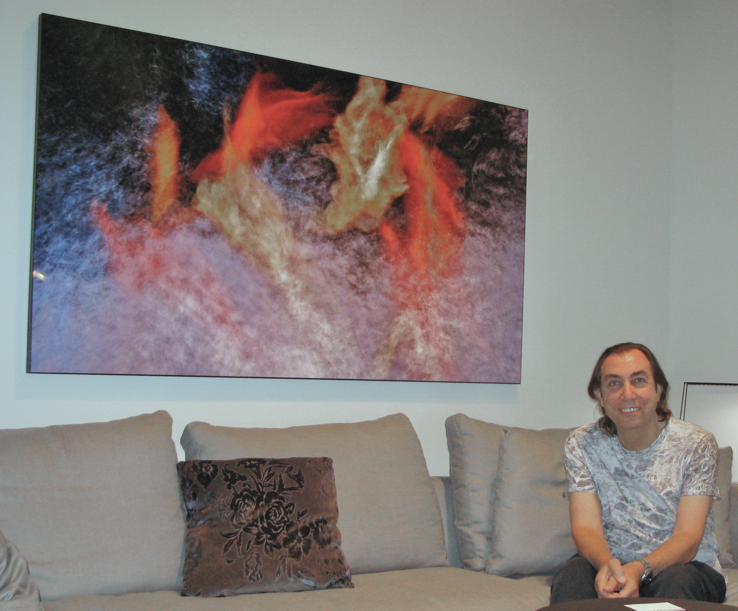 Pen Densham sat next to a print of an abstract, motion-blurred composition of vibrant orange and golden-yellow forms resembling koi fish swimming through textured, lavender and deep blue water.