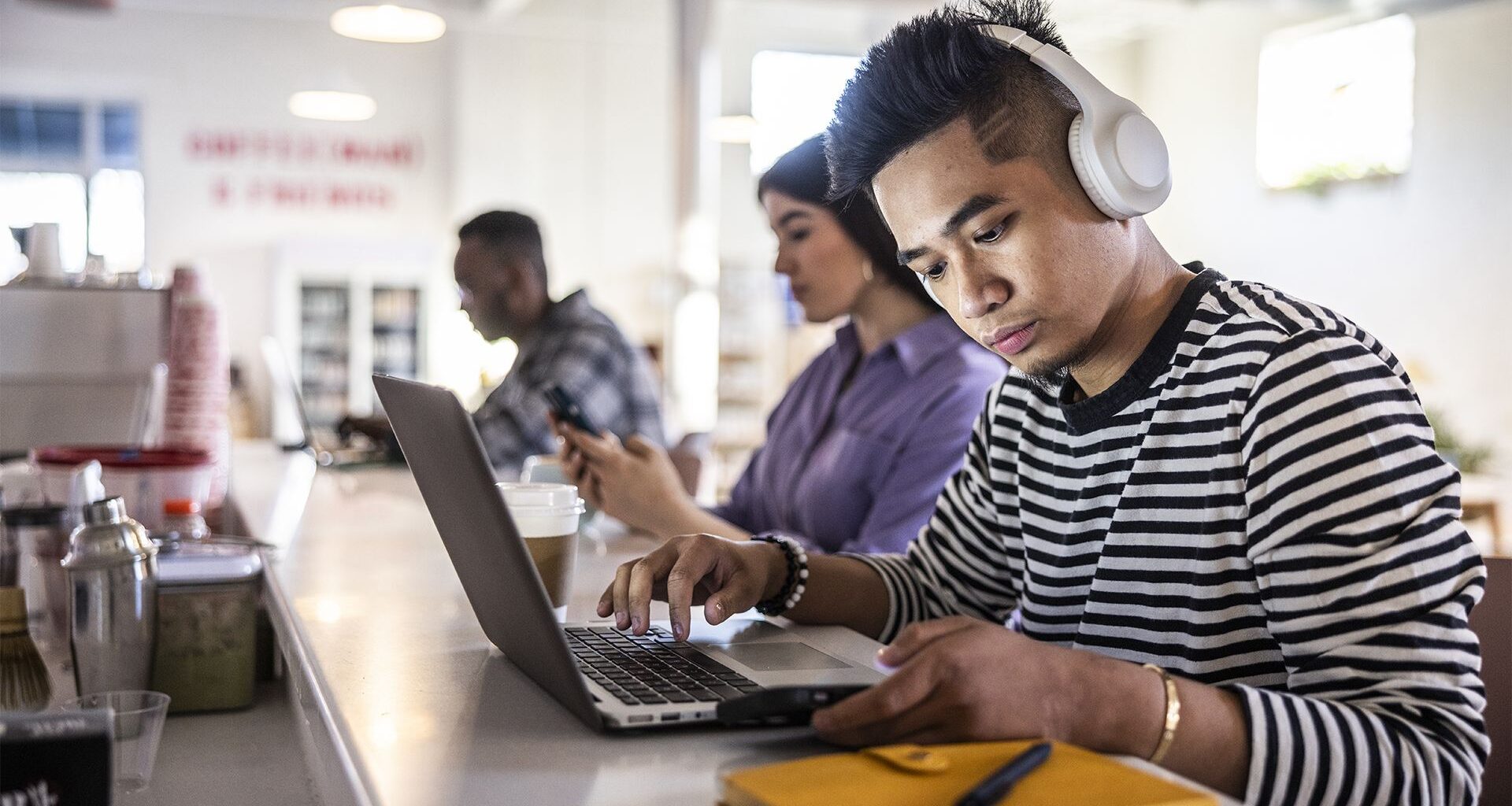 A man sits at the counter top of a coffee shop working on his laptop. In the background, other people are also working. He's wearing headphones to block out distracting sounds to aid concentration