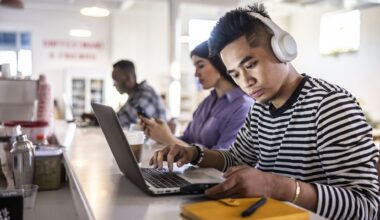A man sits at the counter top of a coffee shop working on his laptop. In the background, other people are also working. He's wearing headphones to block out distracting sounds to aid concentration