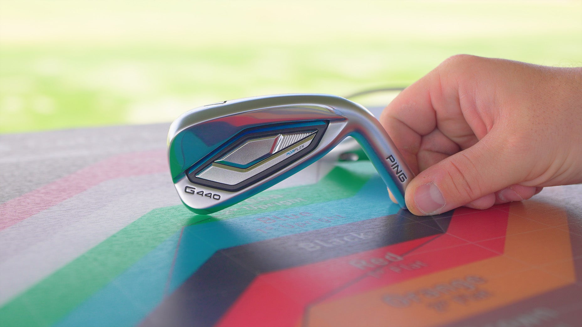 A hand holding a Ping G400 golf club iron rests on a colorful, multi-sectioned chart, with a blurred green outdoor background.