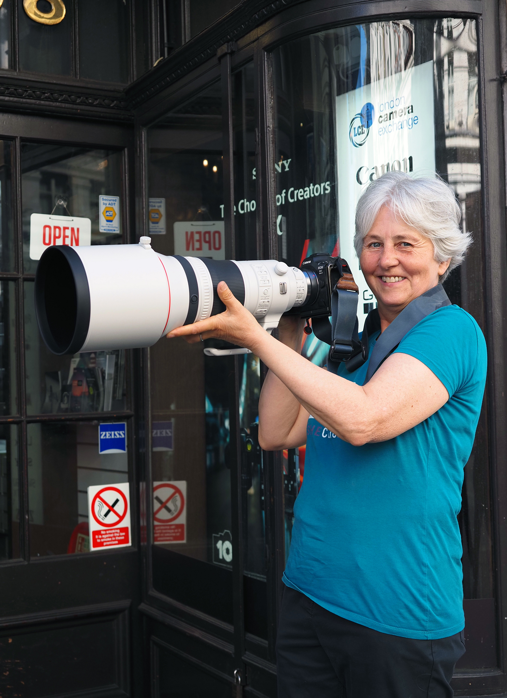 Person with holding a large telephoto camera lens, standing outside a camera store