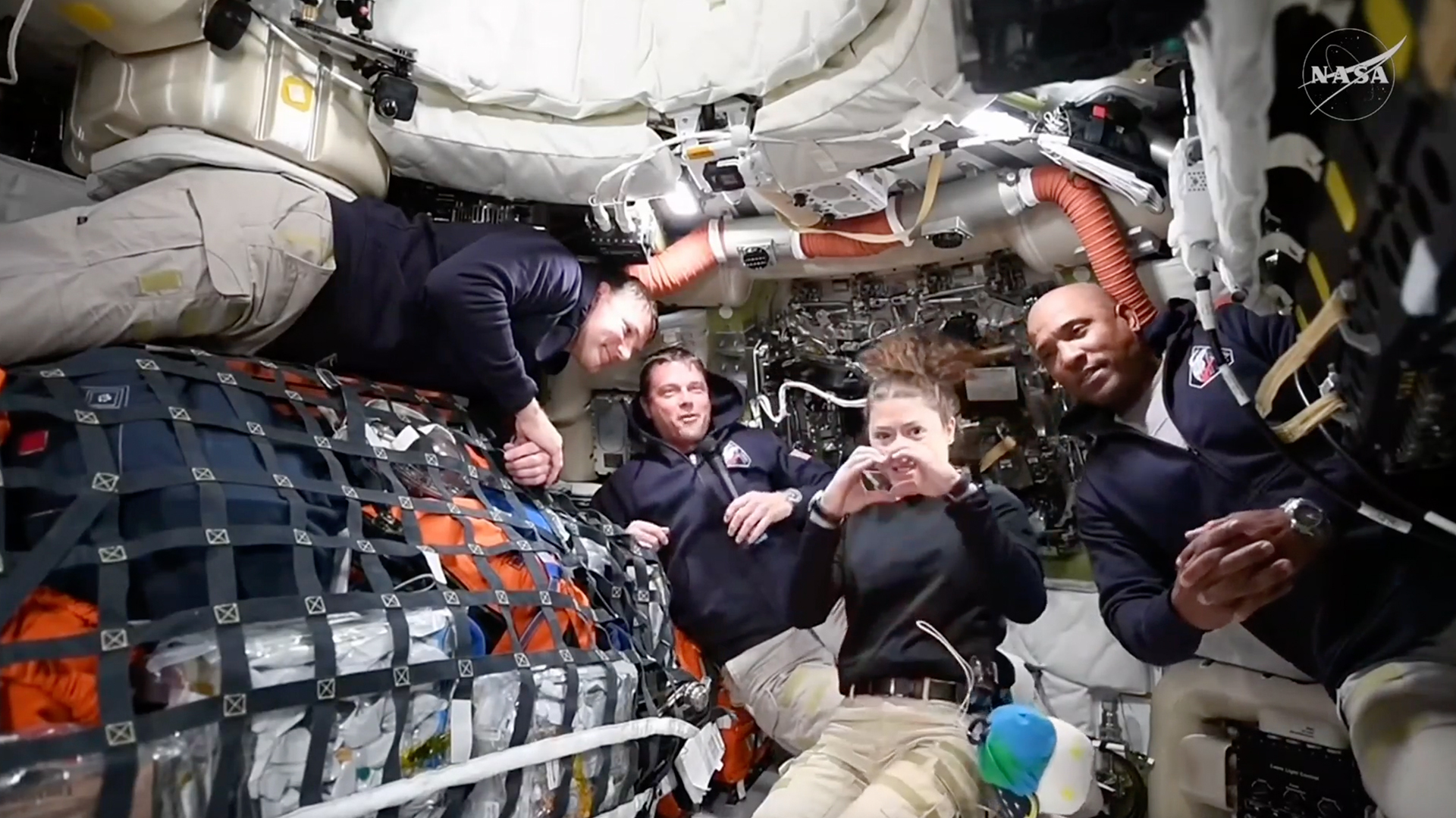 four people in black sweatshirts inside a cramped spacecraft cabin packed with cargo held down by netting and tubes and wires