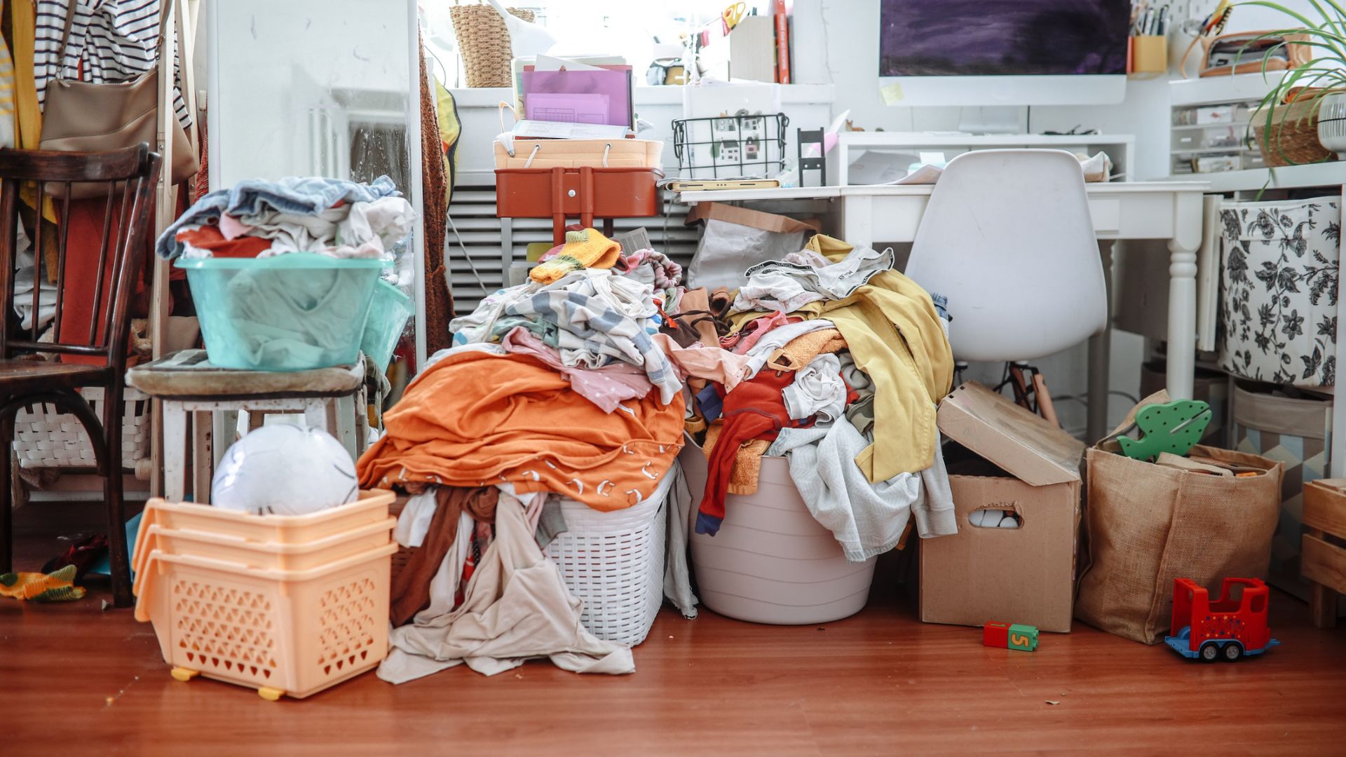 A cluttered bedroom with wood floors showing laundry baskets piled up with clothes and random toys and objects on the desk and floor