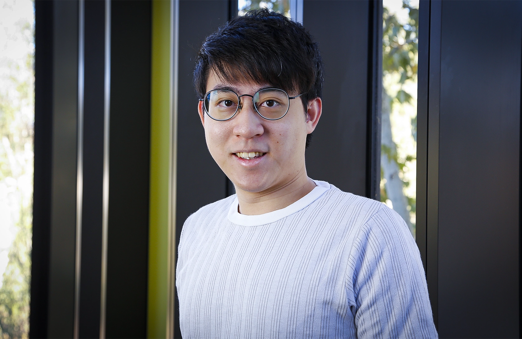 A smiling man wearing round glasses and a light gray ribbed sweater, photographed in front of a dark-framed window.