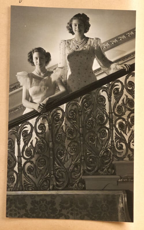 Two women in vintage, formal dresses stand on a staircase with an ornate, decorative railing. One woman stands on a higher step, and both look toward the camera, smiling. Light shines from above, highlighting their faces.