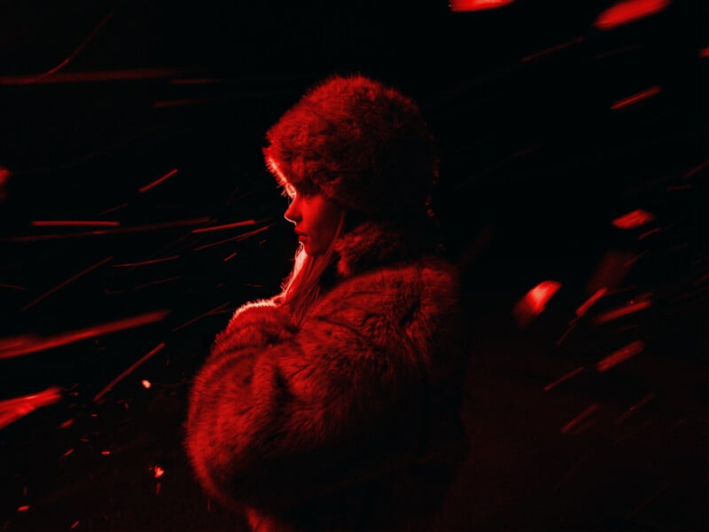 A person in a fur hat and coat stands in profile, surrounded by swirling red light streaks against a dark background, creating a dramatic and mysterious atmosphere.