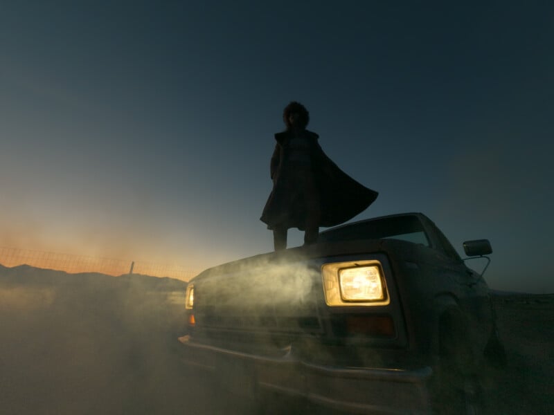 A person stands on the hood of a car with headlights on, surrounded by mist or smoke, silhouetted against a darkening sky at dusk, with mountains faintly visible in the background.