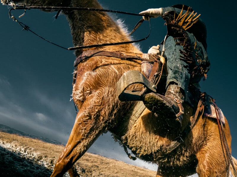 A dramatic, low-angle view of a rider on a galloping, wet horse, with the rider’s boot and stirrup in the foreground and the sky and open landscape in the background.