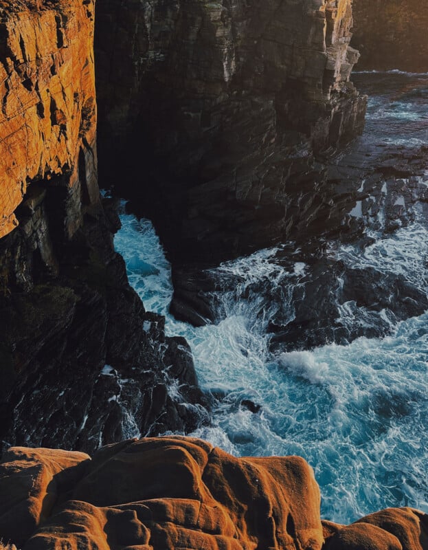 Waves crash against rugged, dark cliffs at sunset, casting warm golden light over the rocks and creating dramatic contrasts between the illuminated and shadowy areas.