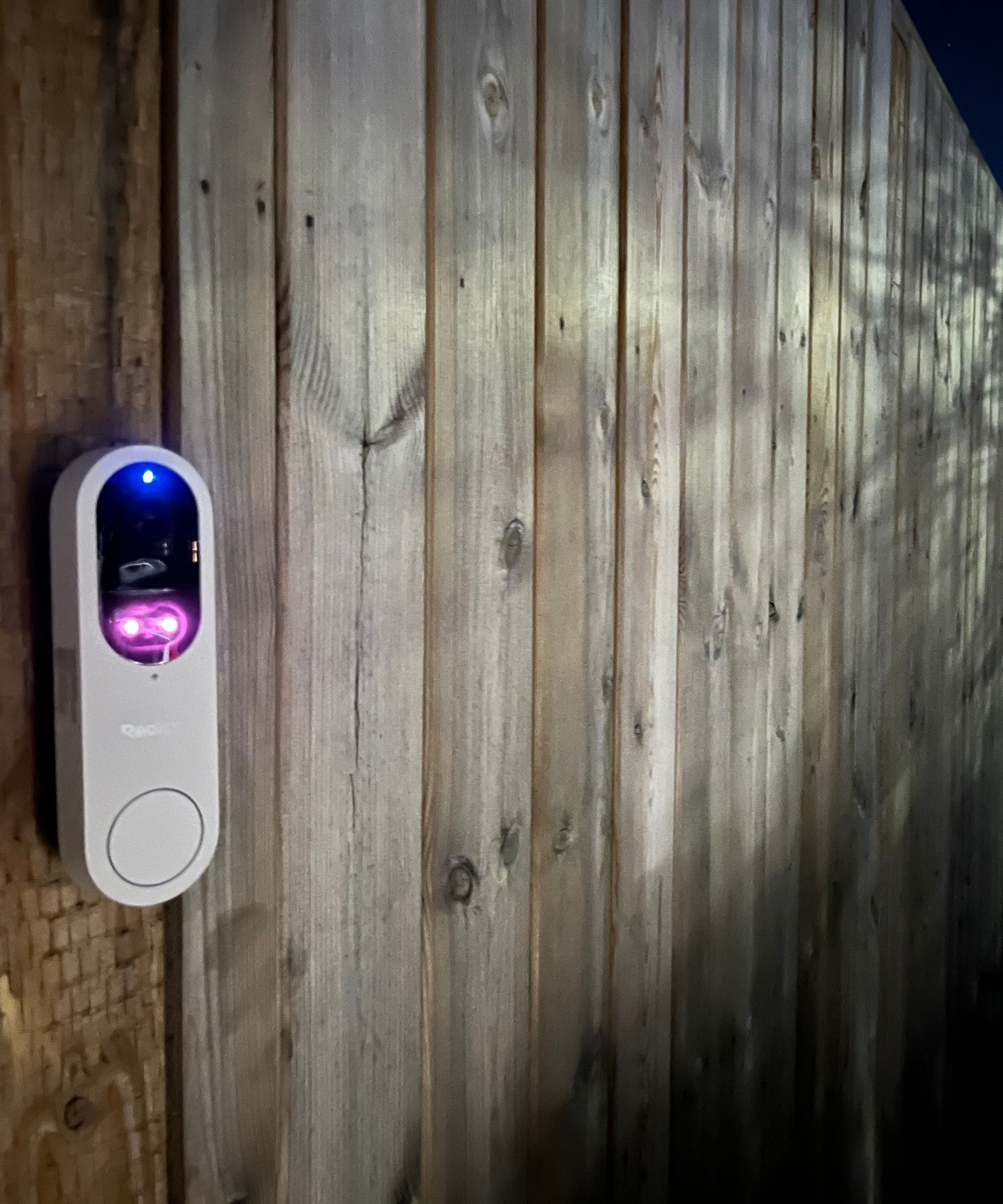 Reolink video doorbell (battery) at nighttime on wooden fence, with lights shining from device.