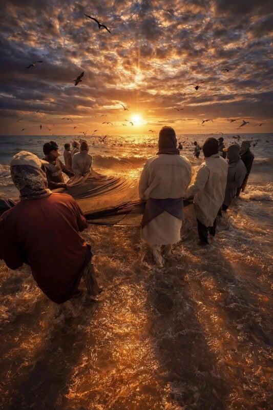 A group of people stands ankle-deep in the ocean at sunrise or sunset, pulling in a fishing net under dramatic, cloudy skies with birds flying overhead. The sunlight reflects warmly on the water’s surface.