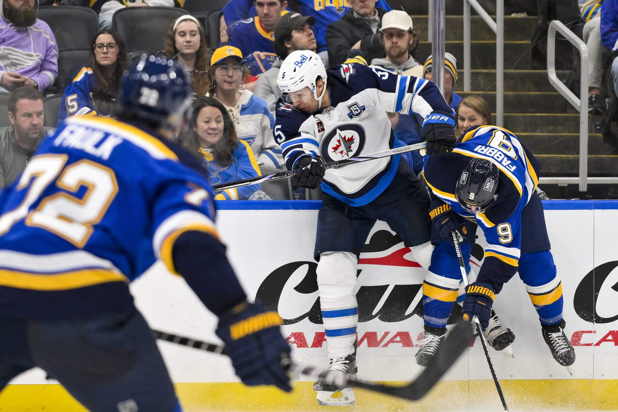 Luke Schenn throws a hit against St. Louis Blues player Robby Fabbri, with another Blues player in the foreground.