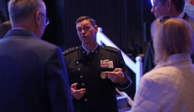 a man in a black military uniform speaks to a group of attendees on a conference ballroom floor