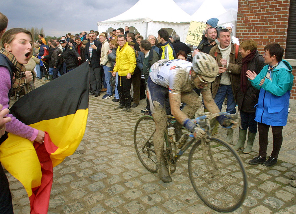 A side shot of Servais Knaven, caked in mud, on a cobbled section of Paris-Roubaix 2001, which is lined by fans