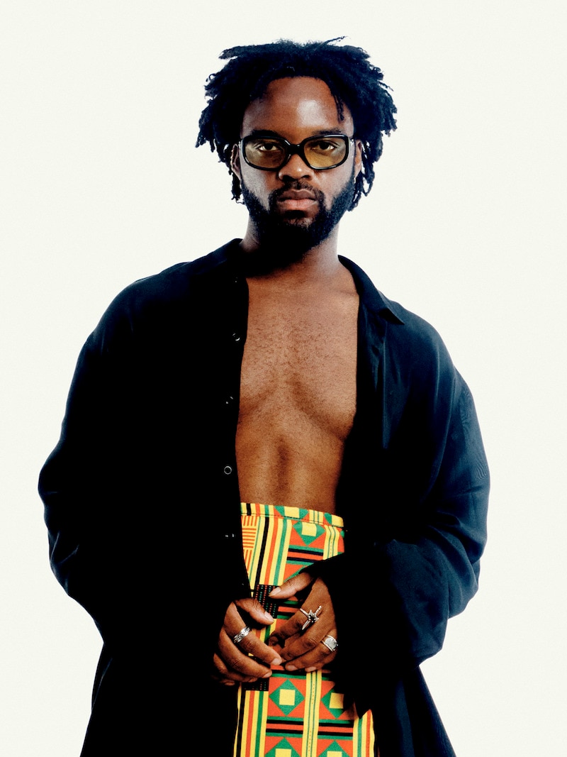Genesis Owusu with bare chest, open black coat and Kente cloth from waist down