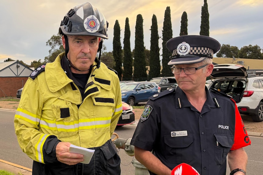 A firefighter and a police officer in front of buildings and trees speaking to media microphones