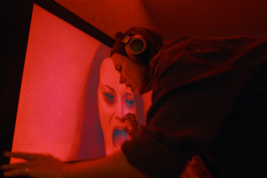 A woman in headphones paws at a red screen with a screaming woman on it