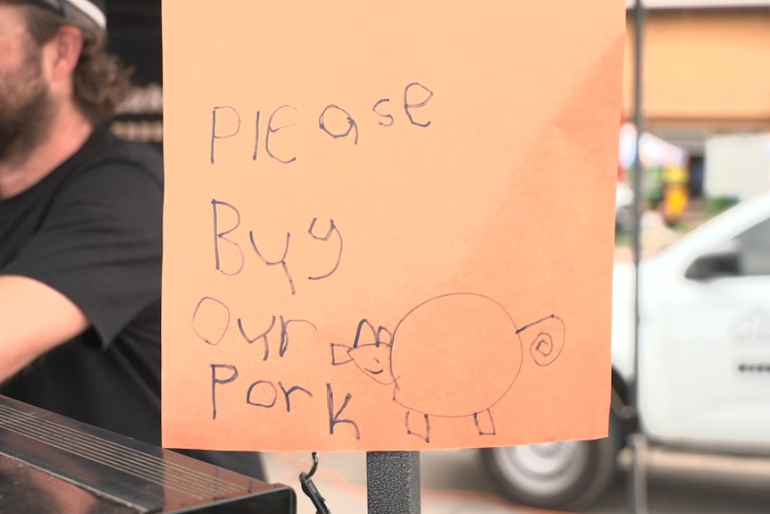 A paper sign with a kid's writing scrawled with the words "please buy our pork" and a cartoon pig attached to a stall awning