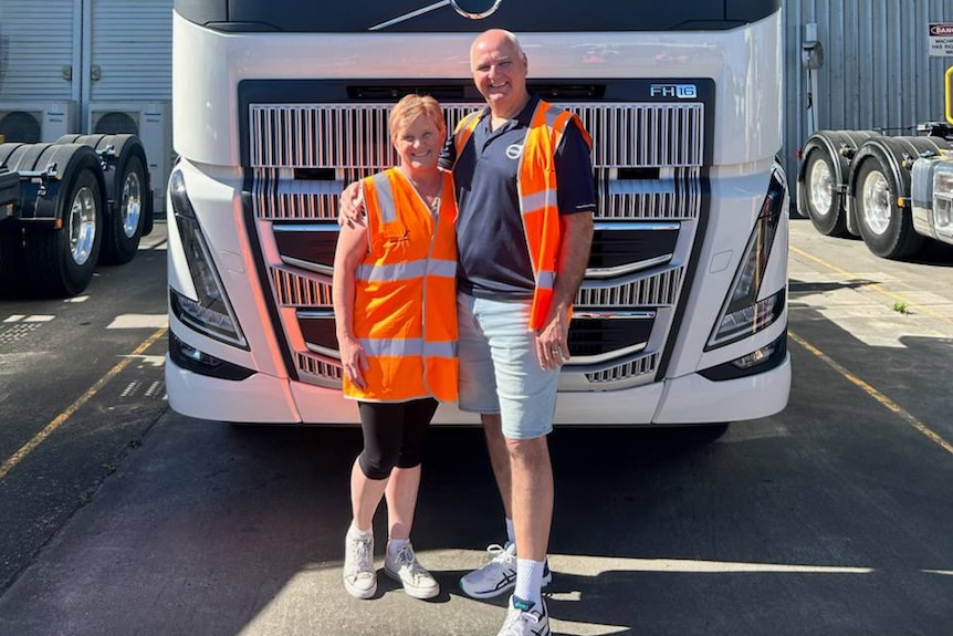 Two people in high visibility vests stand in front of a the grill of a large truck.