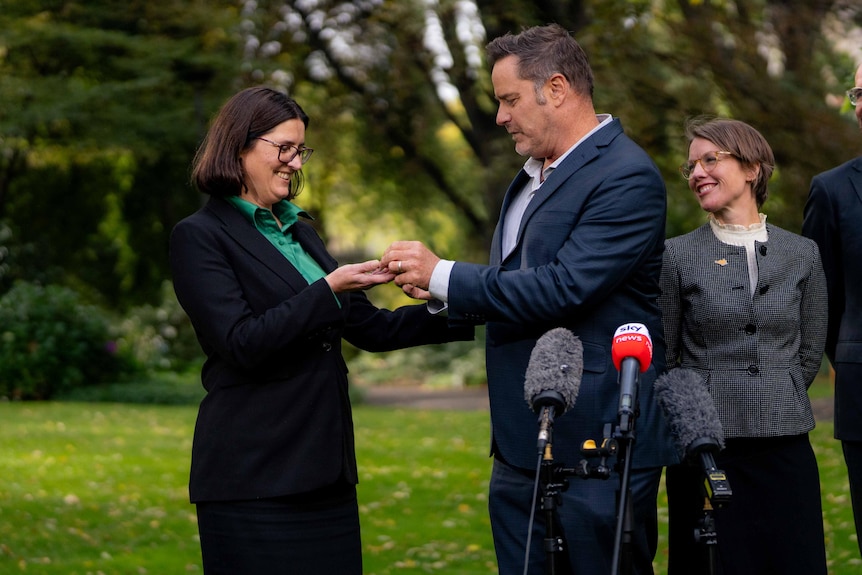 A man in a blue suits hands a small golden pin to a woman with dark brown hair and green shirt.