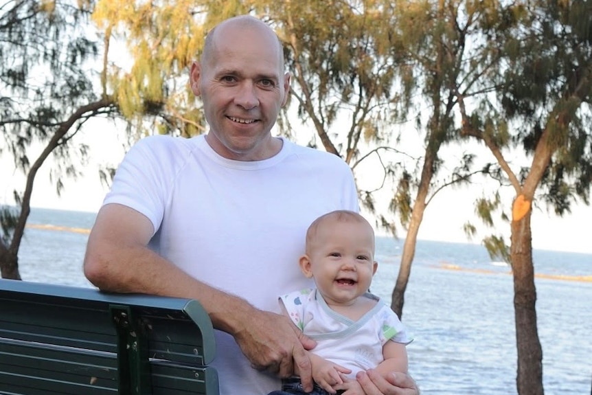 A man sits on a bench by the water with a baby on his knee