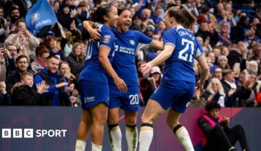 Lauren James celebrates scoring at Stamford Bridge as Sam Kerr and Niamh Charles join her in a huddle, with fans applauding in the stand behind them
