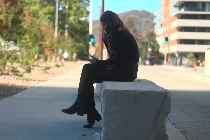 A young woman sits on an outdoor stone bench vaping.